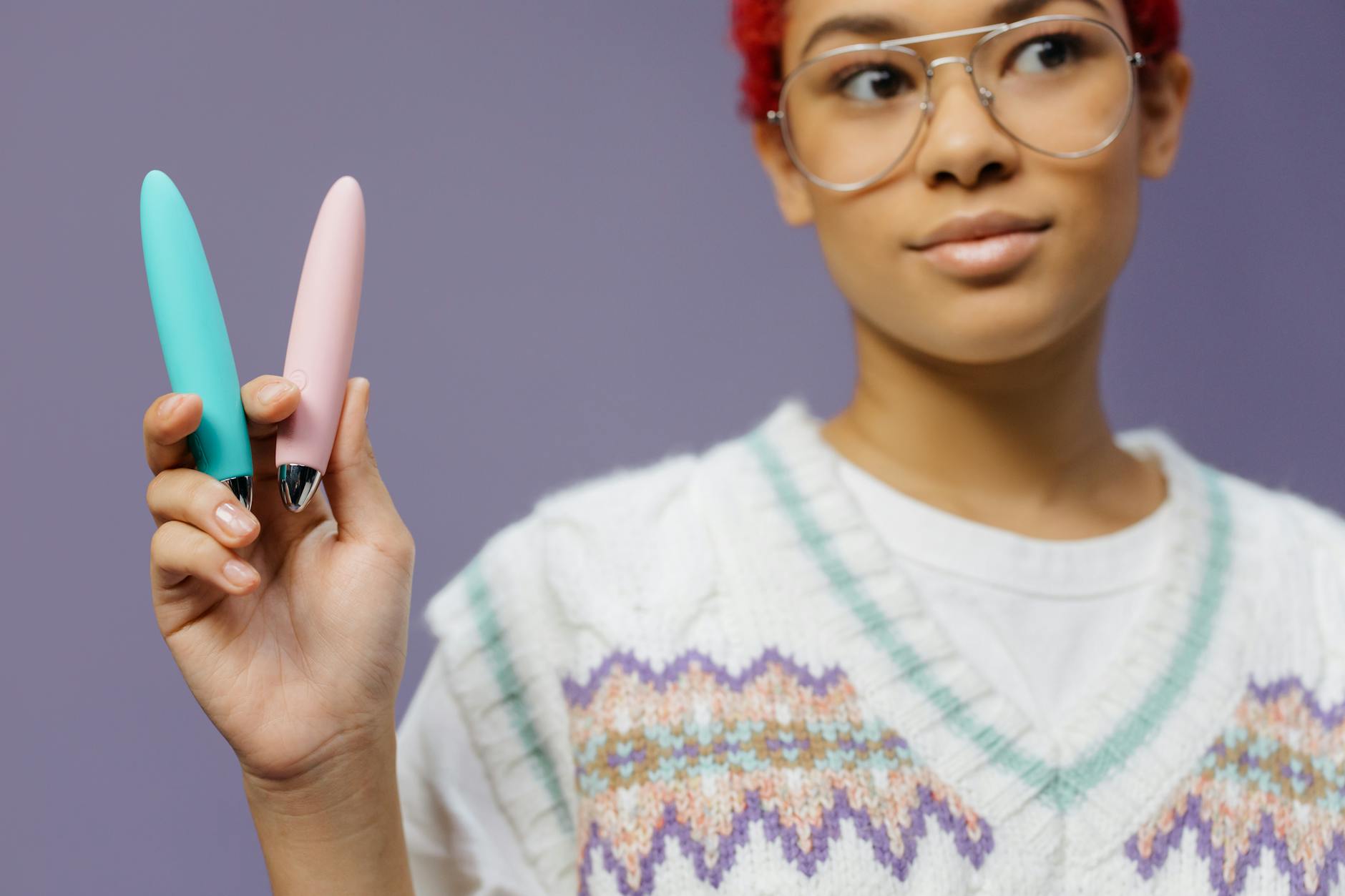 A person with eyeglasses holding two colorful vibrators against a purple background.